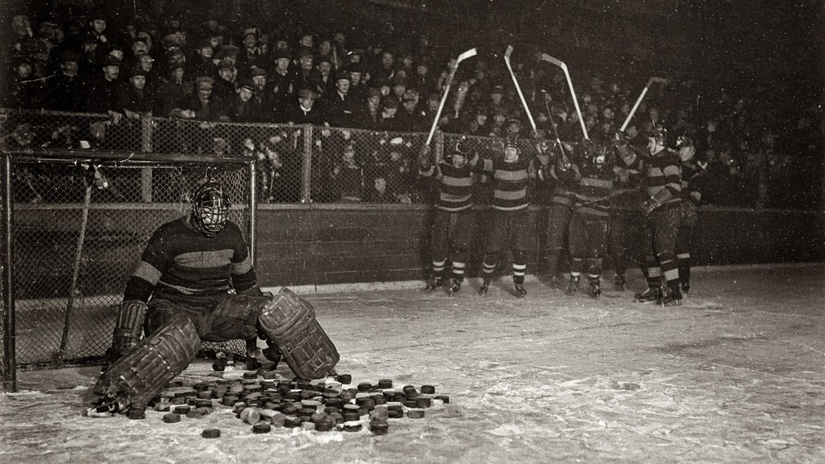 1920s flash photograph of a goaltender in a wire cage mask and leg pads slumped on the ice in his crease among a scattered pile of pucks, while a line of skaters in striped sweaters celebrates with sticks raised in the background